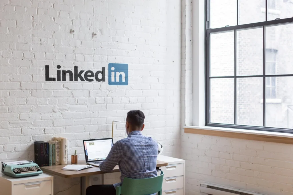 Homem sentado mexendo no notebook com logo do linkedin na parede